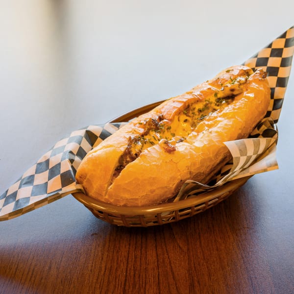 A long, cheesy baked sandwich with herbs, served in a basket with checkered paper on a wooden table.