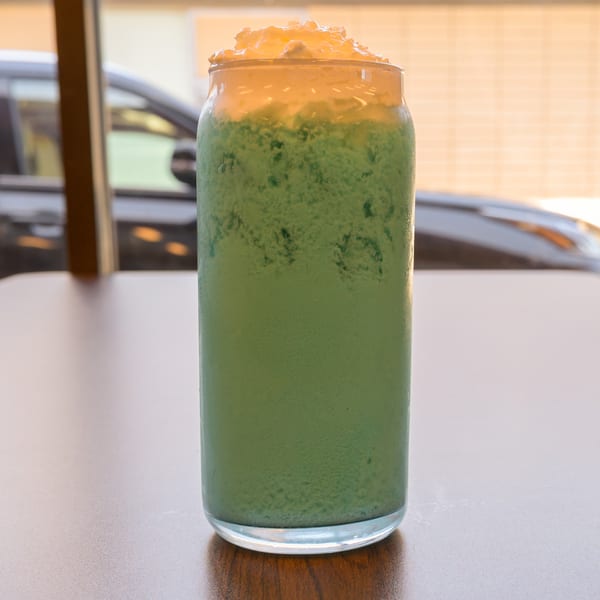 Tall glass of green blended drink with whipped cream on a table, blurred car in background.